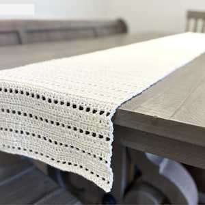 Könnte beinhalten: Cremefarbener, gehäkelter Tischläufer. Der Läufer ist rechteckig und hat ein detailliertes Lochmuster an den Rändern. Er liegt auf einem Holztisch und zeigt seine Textur und sein Design. Die neutrale Farbe und die aufwendige Häkelarbeit verleihen Eleganz.