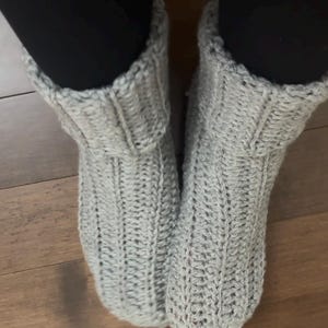 May include: A pair of light gray ankle-length slippers crafted from thick, chunky yarn. The slippers feature a prominent vertical ribbed knit pattern throughout the body and a folded cuff at the top, providing a textured appearance and a snug fit.