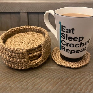 Op de afbeelding: Een set van jute onderzetters en een houder. De ronde onderzetters hebben een geweven textuur. Een witte mok met de tekst "Eat Sleep Crochet Repeat" staat op een van de onderzetters. De set is voor de interieurdecoratie.