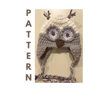 May include: A crocheted owl hat featuring a cream-colored top, gray earflaps, and braided ties. The hat has large circular eyes with dark centers, a brown triangular beak, and small tufts on top. The word PATTERN is printed vertically in brown.