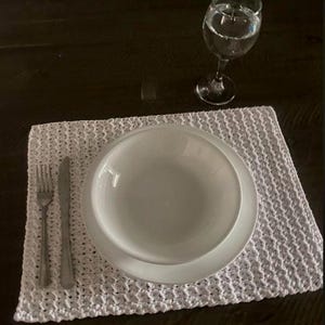 May include: A white crocheted placemat with a white ceramic bowl and a white plate on top of it. A silver fork and knife are placed to the left of the bowl. A clear glass of water is in the background.