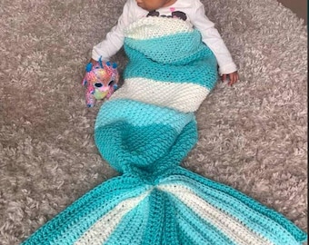 Op de afbeelding: Een turquoise en witte gebreide mermaid-staartdeken. De staart is gestreept en heeft een puntige vin. De deken ligt op een lichtgrijs tapijt.
