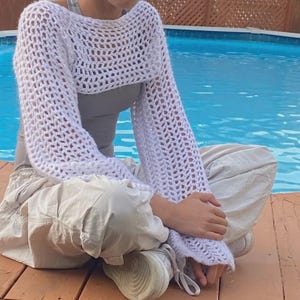 May include: A white, open-weave crochet shrug with long sleeves. The garment is worn over a grey top. The person is sitting on a wooden deck with a swimming pool in the background. The person is wearing beige trousers and white trainers.