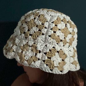 May include: A handmade crochet bucket hat in a granny square pattern. The hat features white and tan yarn, creating a textured accessory. The hat is designed with a brim and a rounded crown. The hat is a stylish accessory.