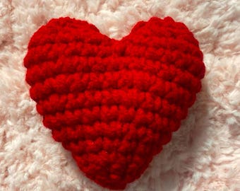 May include: A red crocheted heart-shaped pillow. The pillow is made with a soft, textured yarn and has a slightly bumpy surface.