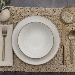Può includere: Una tovaglietta all'uncinetto beige con una ciotola e un piatto in ceramica bianca con bordi marroni. Un tovagliolo di lino beige, una forchetta, un coltello e un cucchiaio in bronzo opaco, un bicchiere a coste e un piccolo vaso bianco.