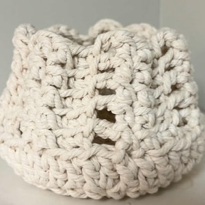 May include: A cream-colored, hand-knitted basket with a rounded base and an open, textured weave. The basket's top edge is gathered, creating a ruffled effect. The thick, natural-toned yarn gives it a rustic, handmade appearance.