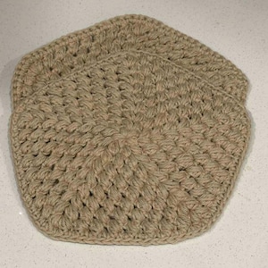 May include: Three beige crocheted coasters are stacked on a white surface. The coasters have a hexagonal shape with rounded corners and a textured, woven appearance. The yarn has a natural, earthy tone.