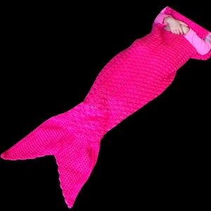 Könnte beinhalten: Eine leuchtend pinkfarbene gehäkelte Meerjungfrauen-Schwanzdecke. Die Decke ist so gestaltet, dass sie einer Meerjungfrauen-Schwanzflosse ähnelt, mit einer spitzen Flosse am Ende.