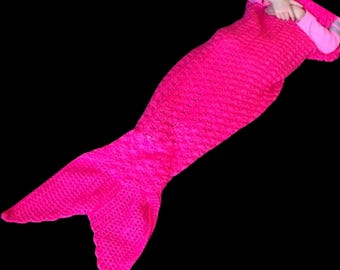 Op de afbeelding: Een felroze gebreide mermaid-staartdeken. De deken is ontworpen om te lijken op een mermaid-staart, met een puntige vin aan het einde.