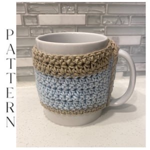 May include: A white ceramic mug with a crocheted cozy. The cozy is made with two colors of yarn: brown and light blue.
