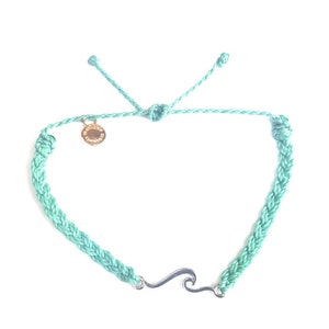 Puede incluir: Un brazalete trenzado turquesa con un dije de plata en forma de ola y un dije de oro. El brazalete es ajustable y se puede usar en la muñeca o el tobillo.