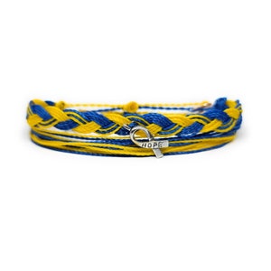 Puede incluir: Un brazalete de amistad trenzado azul y amarillo con un dije de cinta plateado que dice "HOPE".