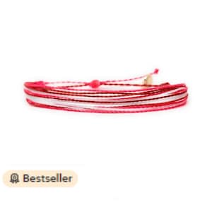 May include: Multi-coloured string bracelet with a pink, white and brown colour scheme. The bracelet is made of thin, braided strings and has a small gold bead at the clasp.