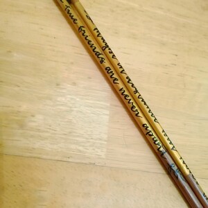 May include: Two wooden drumsticks with dark blue tips. The drumsticks are inscribed with the words "True friends are never apart, maybe in distance but never in heart" in black script. The drumsticks are resting on a light brown wooden surface.