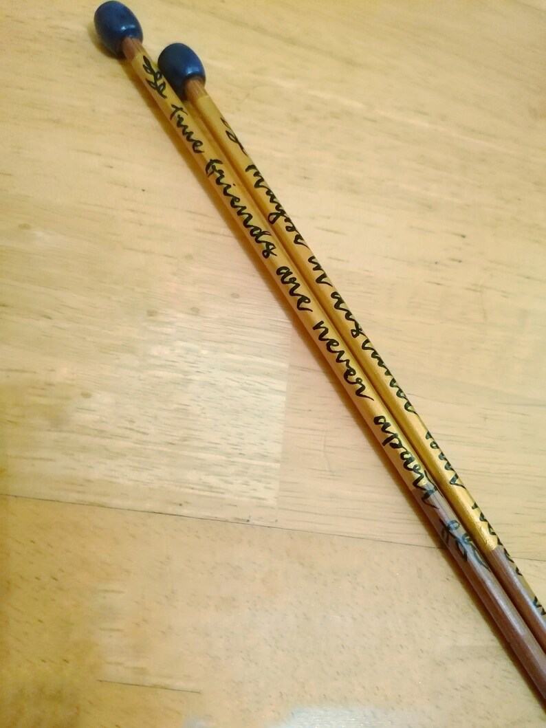 May include: Two wooden drumsticks with dark blue tips. The drumsticks are inscribed with the words "True friends are never apart, maybe in distance but never in heart" in black script. The drumsticks are resting on a light brown wooden surface.
