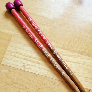 May include: Two wooden knitting needles with red shafts and purple tips. The needles have white script that reads "We love you grandma" and "from Ned and Hugo" with heart accents. The needles are resting on a light wood surface.