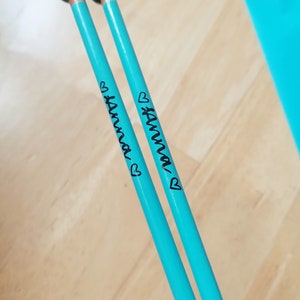 May include: Two turquoise knitting needles with dark green ball tips. The name "Anna" is written in black script on each needle, accompanied by a small heart. The needles are resting on a light wood surface.