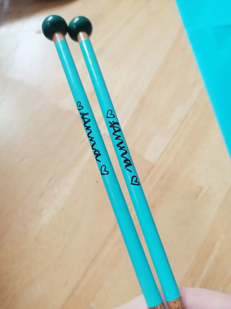 May include: Two turquoise knitting needles with dark green ball tips. The name "Anna" is written in black script on each needle, accompanied by a small heart. The needles are resting on a light wood surface.