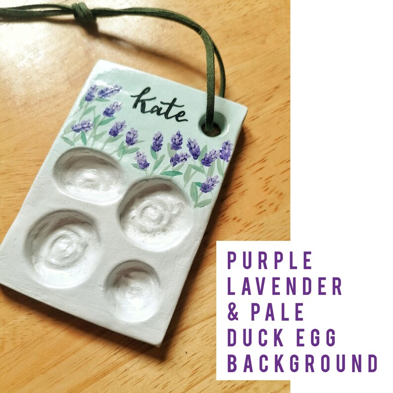 May include: A rectangular, pale green ceramic palette with four circular wells and a lavender floral design. The word "Kate" is written in cursive. A green cord is attached for hanging.