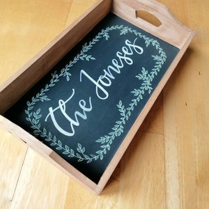 May include: Wooden serving tray with a dark chalkboard centre. The centre features the cursive text "The Joneses" surrounded by a white and green leaf design. The tray has a handle cutout on each end.