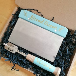 May include: A bread lame and bench scraper set in a gift box. The bench scraper has a light blue handle with the words "Bread Queen" in gold script. The lame has a wooden handle with a light blue accent and the name "Lisa" in gold script.