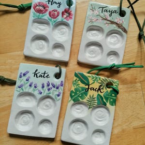 May include: Four rectangular, hand-painted ceramic paint palettes with green cords. Each palette features a different floral design and a name: Amy, Taya, Kate, and Jack. The palettes have six circular wells for holding paint.