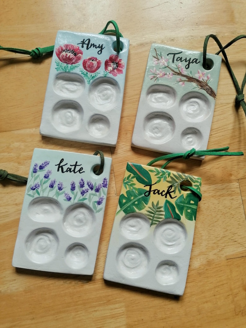 May include: Four rectangular, hand-painted ceramic paint palettes with green cords. Each palette features a different floral design and a name: Amy, Taya, Kate, and Jack. The palettes have six circular wells for holding paint.