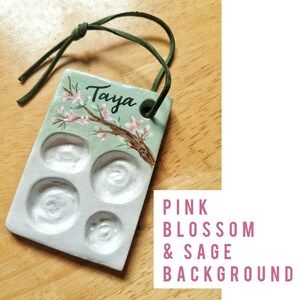 May include: A rectangular, ceramic palette with four wells and a green cord. The palette is painted with a pink blossom branch and the name "Taya". The text "Pink Blossom & Sage Background" is in the lower right corner.
