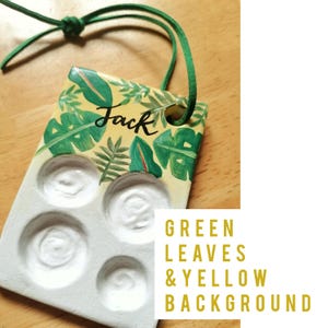 May include: A handmade paint palette with a yellow background and green leaf design. The word "Jack" is written in black script. The palette has four circular wells and a green cord for hanging.
