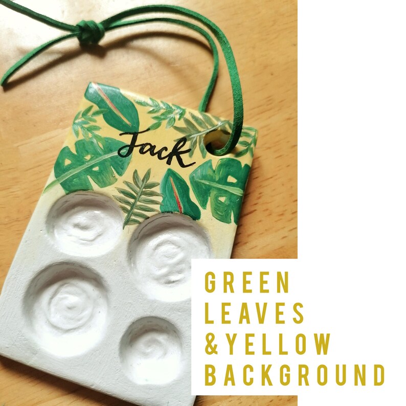 May include: A handmade paint palette with a yellow background and green leaf design. The word "Jack" is written in black script. The palette has four circular wells and a green cord for hanging.