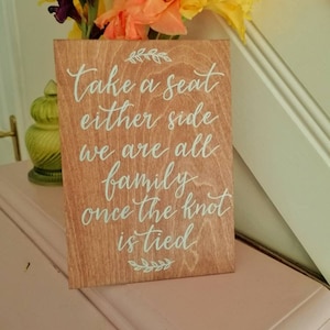 May include: A wooden sign with the text "Take a seat either side we are all family once the knot is tied" in white script. The sign is light brown and is decorated with floral accents. The sign is placed on a pink surface.