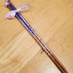 May include: A pair of knitting needles with purple shafts and light brown tips, tied together with a light purple ribbon. The needles are inscribed with the words "Nanna's Knitting Needles" in white script.