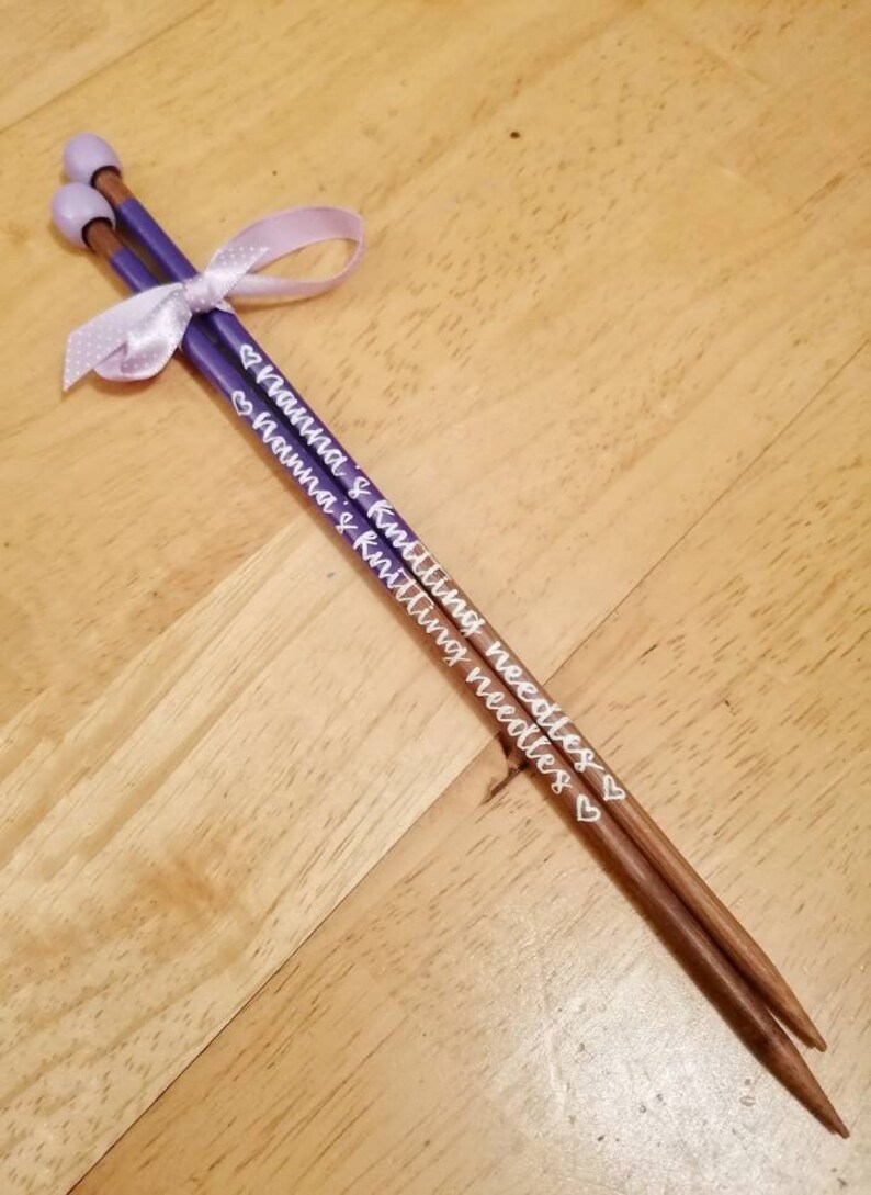May include: A pair of knitting needles with purple shafts and light brown tips, tied together with a light purple ribbon. The needles are inscribed with the words "Nanna's Knitting Needles" in white script.