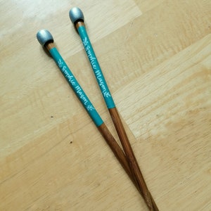 May include: Two wooden chopsticks with teal bands featuring the name "Sophie Mason" in white script and a leaf design. The tips are brown, and the ends are painted silver. The chopsticks are resting on a light-colored wooden surface.