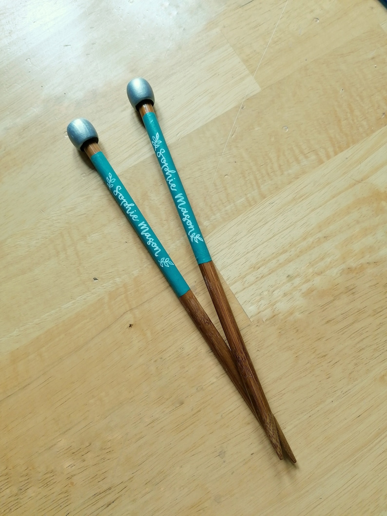 May include: Two wooden chopsticks with teal bands featuring the name "Sophie Mason" in white script and a leaf design. The tips are brown, and the ends are painted silver. The chopsticks are resting on a light-colored wooden surface.