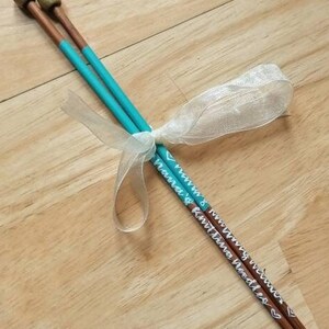 May include: A pair of wooden knitting needles with teal painted shafts and gold-colored tips. The needles are tied together with a white ribbon. The text "Knitting Needles" is written on the needles.