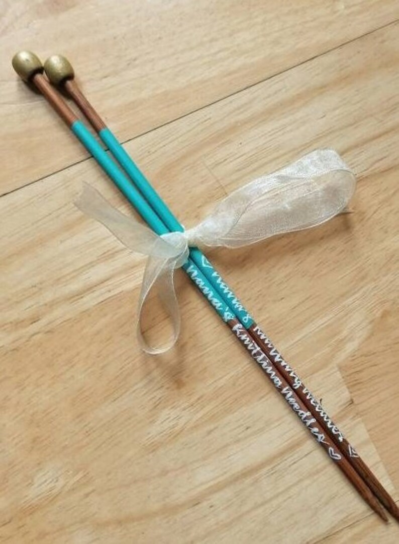 May include: A pair of wooden knitting needles with teal painted shafts and gold-colored tips. The needles are tied together with a white ribbon. The text "Knitting Needles" is written on the needles.