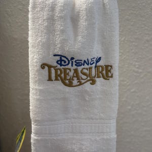 May include: White, rectangular hand towel with the embroidered text "Disney TREASURE" in blue and gold. The towel is hanging on a silver towel ring. The towel has a textured surface and a decorative border.