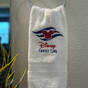 May include: White hand towel with the Disney Cruise Line logo embroidered in red and blue. The logo features Mickey Mouse ears and stylized waves. The towel is hanging on a silver towel ring.