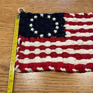 May include: A woven textile flag featuring a dark blue canton with white star-shaped embellishments and alternating red and white horizontal stripes. A yellow measuring tape is positioned along the left edge, showing a height of 8 inches.