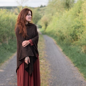 Op de afbeelding: Een vrouw draagt een bruine gebreide poncho met een rode rok en staat op een pad in een landelijke omgeving.