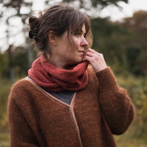 Peut inclure: Une femme porte une écharpe infinie rouille et un cardigan marron. Le cardigan a un col en V et une fermeture boutonnée. L'écharpe est enroulée autour du cou, et la femme regarde sur le côté. L'arrière-plan est flou avec des tons verts et bruns.