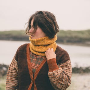 Peut inclure: Un col roulé en tricot jaune moutarde. Le col est porté sur une chemise à carreaux et un cardigan marron avec des bordures orange et des manches à motifs. L'arrière-plan montre un plan d'eau et un ciel nuageux.