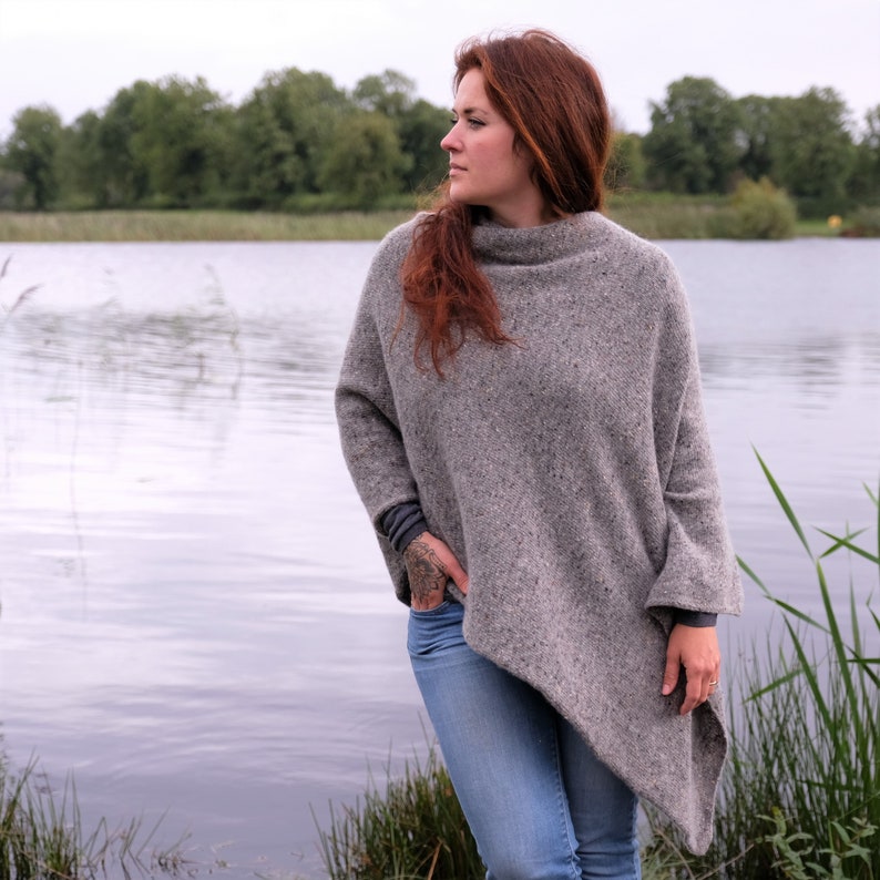 Op de afbeelding: Een vrouw die een grijs, gevlekt, oversized poncho draagt ​​staat aan de rand van een meer. Ze draagt ​​een blauwe jeans en heeft lang, rood haar.