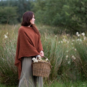 Op de afbeelding: Een vrouw in een roestkleurige poncho staat in een veld met hoog gras en witte wilde bloemen. Ze houdt een rieten mand vol witte bloemen vast.