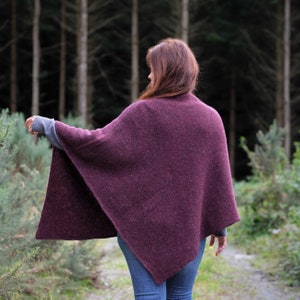 Op de afbeelding: Een vrouw draagt een diep paarse gebreide poncho in een bosrijke omgeving.