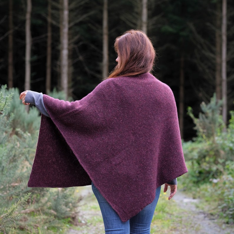 Op de afbeelding: Een vrouw draagt een diep paarse gebreide poncho in een bosrijke omgeving.