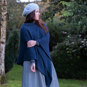 Op de afbeelding: Een vrouw draagt een blauwe gebreide poncho met een grijze baret. De poncho heeft een lang, vloeiend ontwerp en wordt gedragen over een grijs en wit geruite rok.