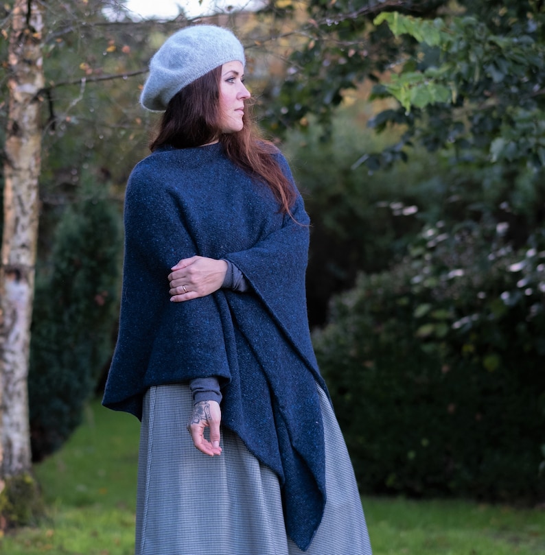 Op de afbeelding: Een vrouw draagt een blauwe gebreide poncho met een grijze baret. De poncho heeft een lang, vloeiend ontwerp en wordt gedragen over een grijs en wit geruite rok.